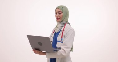 Beyaz arka planda tesettürlü Müslüman kadın doktor dizüstü bilgisayar tutuyor. Dost canlısı sıhhiye dalgaları hastaya çevrimiçi randevuda el sallıyor