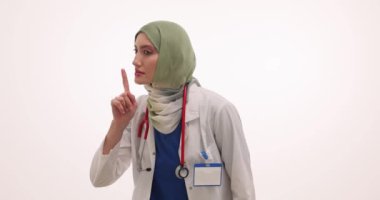 Başörtülü kadın doktor beyaz arka planda sessizlik jesti yapıyor. Üniformalı Müslüman çocuk doktoru genç hastalara hastanede sessiz olmalarını söyledi