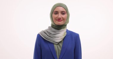 Hoş gülüşü olan Müslüman genç bir kadın stüdyoda beyaz arka planda fotoğraf için poz veriyor. Tesettürlü ve mavi ceketli neşeli bir yüz ifadesi olan bayan.