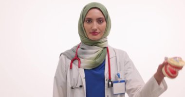 Başörtülü kadın doktor beyaz arka planda çene modeli ve diş fırçası gösteriyor. Müslüman kadın diş hekimi hastaya diş temizliğini açıklıyor