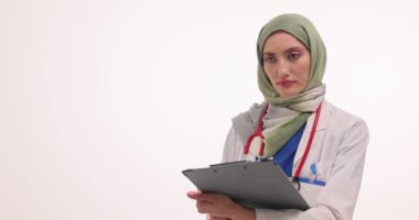 Başı beyaz arka planda olan tesettürlü kadın doktor başını sallıyor. Üniformalı Müslüman kadın sağlık alanında profesyonelliği yansıtıyor