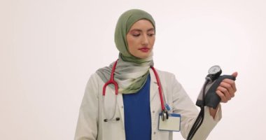Müslüman kadın aile doktoru beyaz arka planda tansiyon monitörü gösteriyor. Türbanlı kadın terapist, cihazın hastaya kullanımını açıklıyor