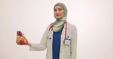 Üniformalı kadın doktor kalp modelini gösteriyor. Neşeli yüz ifadesi olan Müslüman kardiyolog, yaşlı hastaya hastalığı açıklıyor