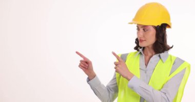 Beyaz arka planda işaret parmakları olan miğfer başlıklı kadın mühendis. Koruyucu üniformalı genç bir kadın aktif jestler kullanarak dikkatini veriyor.