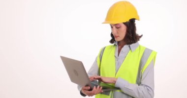 Beyaz duvara karşı dizüstü bilgisayarda odaklanmış kadın mühendis tipleri. Miğfer ve yelek giyen kadın işçi inşaat alanını yavaş çekimde kontrol ettikten sonra rapor oluşturur
