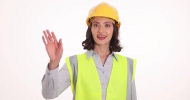 Beyaz arka planda güvenlik yeleği ve kask dalgaları olan kadın mühendis. Mutlu yüz ifadesi olan üniformalı bir kadın iş arkadaşlarını ağır çekimde selamlıyor.