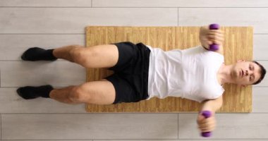 Atletik genç adam evde paspasın üzerinde iki dambılla egzersiz yapıyor ve kaldırma hareketleri yapıyor. Sağlık ve Spor