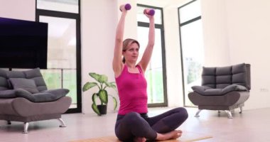 Kadın kilo veriyor ve sabah evde yoga minderinde oturan halterlerle egzersiz yapıyor. Sağlıklı yaşam tarzı ve eğitim