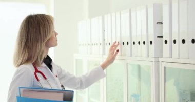 Doktor raftan yüzüklü beyaz klasörü alır. Tıbbi üniformalı bir kadın klinik odasında arşiv malzemesi arıyor.