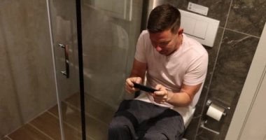 Erkek oyuncu evdeki tuvalette akıllı telefondan mobil oyun oynuyor. Çekici adam mutlu olur ve telefonla rahatlar.
