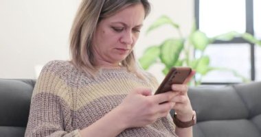 Kadın telefonda koltukta oturan üzücü mesajları okuyor. Bayan endişeli ifadesiyle sosyal medyada bilgi kaydırırken endişeli görünüyor.