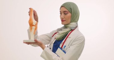 Başörtülü kadın doktor beyaz arka planda diz eklemi modeli gösteriyor. Üniformalı Müslüman kadın fizyoterapist konferansta eklemlerin çalışmalarını gösteriyor