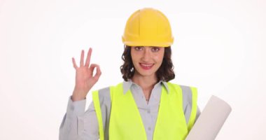 Güvenlik yeleği ve miğferi olan kadın mühendis, iyi bir jest olduğunu gösteren planlar tutuyor. Gülümseyen kadın inşaat sektöründeki kariyerinden memnuniyetini dile getiriyor