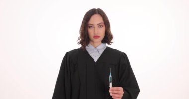 Siyah elbiseli kadın yargıç elinde beyaz arka planda kelepçe gösteren bir şırınga tutuyor. Kadın, hukuk sistemi ve adalet kavramında otoriteyi somutlaştırıyor