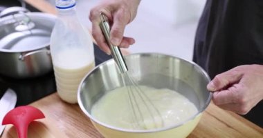 Adam kahvaltı hazırlıyor, metal kasede sıvı malzemeler çırpıyor. Erkek aşçı ahşap tezgahta duruyor krep için süt ve hamur malzemelerini karıştırıyor.