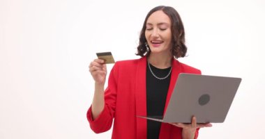 Canlı kırmızı blazer ceket giyen iş kadını dizüstü bilgisayar kullanarak online alışveriş yapıyor. Esmer kadın beyaz arka planda kredi kartı ve laptop tutuyor.