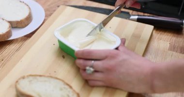 Kadın plastik konteynırdan taze ekmeğe krem peynir sürüyor. Ev hanımı ağır çekimde yemek için tahtanın üzerine yayılmış sandviç yapar.