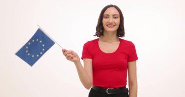 Genç kadın gülümsüyor ve beyaz arka planda Avrupa Birliği bayrağı sallıyor. Kadın uluslararası işbirliğini destekliyor ve ağır çekimde seyahat etmeyi hayal ediyor