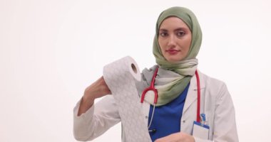 Müslüman kadın doktor tuvalet kağıdını rulo kağıttan söküyor. Steteskopla tesettürlü gülümseyen kadın doktor tuvalet kağıdını beyaz duvara yapıştırıyor.