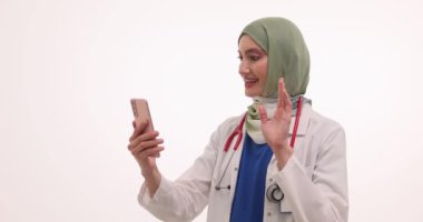 Tesettürlü kadın doktor el kol hareketi yapıyor. Laboratuvar önlüklü kadın doktor. Akıllı telefondaki video görüşmesi sırasında el sallıyor.