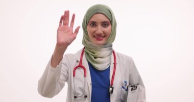 Tesettürlü genç bayan doktor gülümseyerek el sallıyor. Steteskoplu Müslüman kadın doktor hastaları karşılama jesti yapıyor.