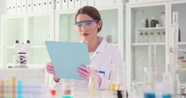 Laboratuvar önlüğü ve güvenlik gözlüğü takan kadın bilim adamı incelemeleri titizlikle laboratuvarda rapor veriyor. Kadının elinde enstrüman ve ekipman panosu var.