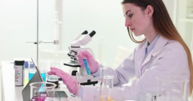 Kadın bilim adamı, iş yerindeki tablette mikroskopla test tüpü girdi sonuçlarını tutuyor. Üniformalı gevşek saçlı laboratuvar çalışanı çalışma sürecine dahil oluyor.
