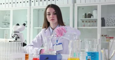 Kadın bilim adamı laboratuvarda terazinin üzerine şişeden cama sıvı döküyor. Üniformalı teknisyen kimyasal deney için reaktörler hazırlıyor.