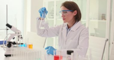 Güvenlik gözlüklü esmer bilim adamı laboratuarda sıvı incelerken şişe sallıyor. Kadın teknisyen deney sırasında kimyasal reaksiyonları kontrol ediyor.