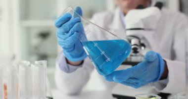 Bilimadamı Erlenmeyer şişesini parlak mavi sıvıyla doldurmuş. Kadın masadaki kimyasal reaksiyonlardan sonra çözümü mikroskopla inceliyor.
