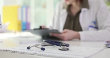 Kadın doktor akıllı tabletteki tıbbi kayıtları inceliyor. Steteskop, teknolojinin sağlık sektörüne entegrasyonunu ön plana çıkaran masa başında yatıyor
