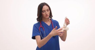 Mavi önlüklü kadın doktor, beyaz üzerindeki yapıyı açıklayan ayak anatomisi modelini tutuyor. Steteskoplu kadın doktor açıklama getiriyor