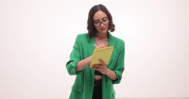 Yeşil ceketli odaklanmış iş kadını beyaz arka plan üzerine not defteri tutuyor. Düşünceli kadın, bir aylık çalışma programı oluşturuyor.