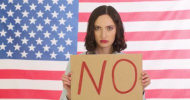 Kararlı ve ciddi bir kadın güçlü duruyor, Amerikan bayrağının önünde belirgin bir HAYIR işareti taşıyor.