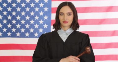 Cüppeli kendinden emin bir kadın yargıç, Amerikan bayrağına karşı tokmak çapraz kollar tutuyor. Gülümseyen kadın, kariyerinin yavaş ilerlemesiyle tatmin olur.