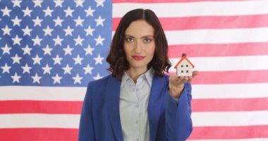 Mutlu kadın emlakçı Amerikan bayrağına karşı anahtar ve küçük oyuncak evi tutuyor. Resmi kıyafet içinde gülümseyen genç bir kadın ev almayı teklif ediyor.
