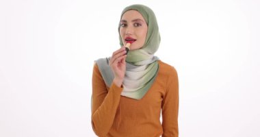 Tesettürlü genç bir kadın beyaz arka planda buluşmaya hazırlanırken kırmızı ruj sürüyor. Gülümseyen Müslüman kadın güzellik rutinini yavaş çekimde yapar.