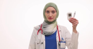 Tesettürlü Müslüman kadın doktor elinde bir bardak şeker küpü ve glukometre cihazı tutuyor. Bayan karbonhidrat seviyelerini ve vücut durumunu yavaş çekimde izlemeyi tavsiye ediyor.