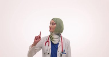 Türbanlı Müslüman kadın doktor işaret parmağıyla işaret ediyor ve yukarı doğru uyarıda bulunuyor. Steteskoplu bir kadın ağır çekimde iş yapma konusunda kendine güveniyor.