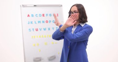 Resmi takım elbiseli kadın öğretmen beyaz tahtaya dur işareti gösteriyor. Dişi pedagog öğrencilerden ders sırasında konuşmayı kesmelerini istiyor