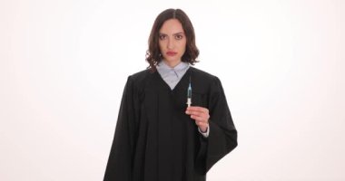 Esmer kadın yargıç beyaz arka planda şırınga ve kelepçe gösteriyor. Siyah elbiseli avukat ceza davasında nihai karara hazırlanıyor.