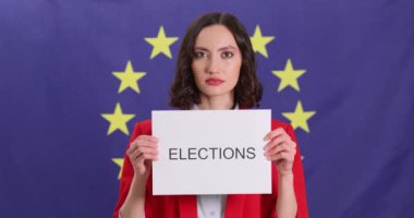 Ciddi ifadeleri olan bir kadın, arka planda Avrupa Birliği bayrağı olan ELECTION sözcüğüyle işaret dili gösteriyor. Kadın demokratik süreçlere destek gösteriyor