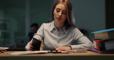 Kadın ofis çalışanı ofiste geç saatlere kadar çalışırken yorgun hissediyor. Yorgun iş kadını, iş yerinde akıllı telefondan saate bakarken esniyor.