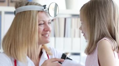 Kadın doktor otoskop kullanan bir kızın kulağını muayene ediyor. Yansıtıcı bantlı kadın otolarolog kulak enfeksiyonu belirtilerini kontrol ediyor.