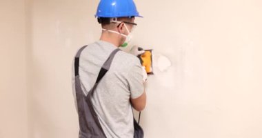 İnşaat işçisi, duvar temizleme işine girişiyor. Miğfer ve koruyucu yüz maskesi takan deneyimli bir erkek yenileme çalışması yapıyor.