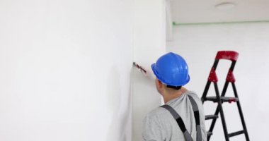 Miğferli adam merdiven karşısında beyaz renk kullanarak duvarı fırçayla boyuyor. Üniformalı dikkatli inşaat işçisi yenileme sürecine giriyor