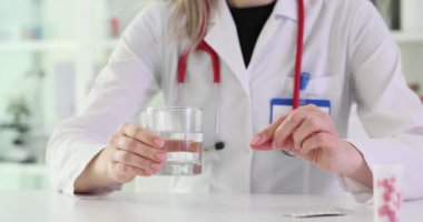 Kadın doktor, hastane işyerindeki masada su bardağı ve hap gösteriyor. Terapist klinikte içkiyle birlikte eczane almayı öneriyor.