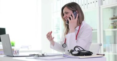 Kadın doktor klinikte masa başında çalışırken telefonla konuşuyor. Hasta kaydını dolduran kadın hekim randevuda cep telefonu sohbetine girişiyor