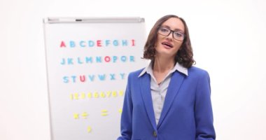 Mutlu öğretmen ilgiye minnet duyarak selamlıyor. Kadın İngilizce öğretmeni, yavaş çekimde mıknatıslı harflerle tahtada tiyatro hareketi gösteriyor.