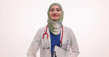 Tesettürlü neşeli kadın doktor beyaz arka planda el yazısı tutuyor. Müslüman kadın sıhhiyeci hastayı hastane randevusunda karşıladı
